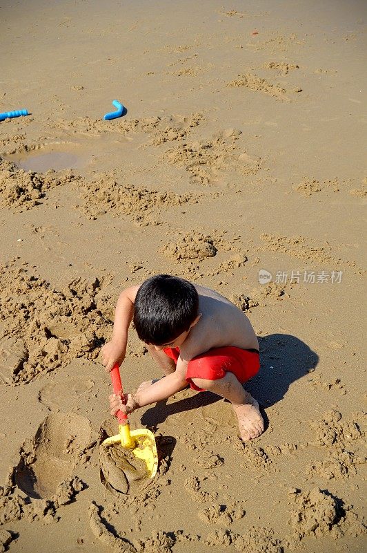 一个穿着红色短裤的小孩坐在沙滩上玩玩具，拿着铲子在沙滩上挖沙子