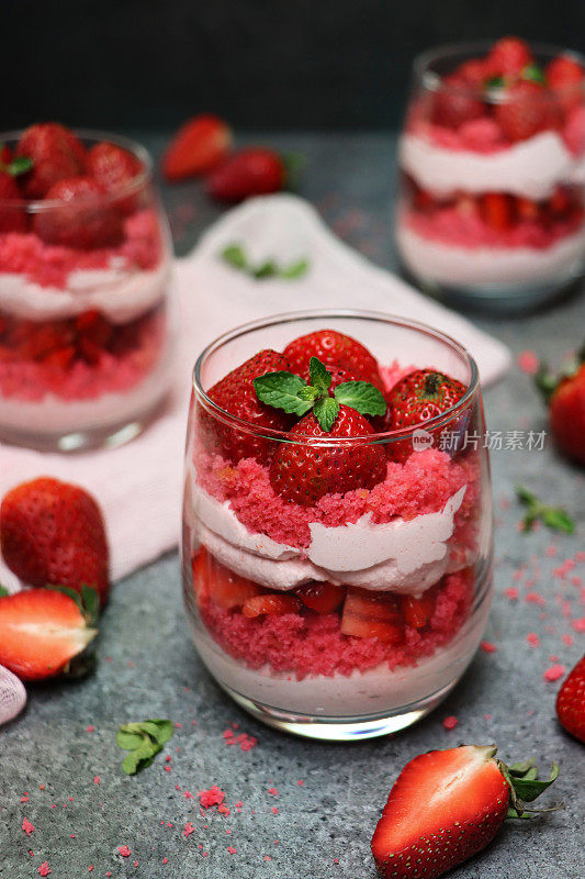 图像眼镜包含自制的草莓慕斯甜点，上面覆盖着整个草莓，奶油慕斯和红丝绒蛋糕屑的分层布丁，周围是蛋糕屑和水果，灰色的表面，高架的视野，聚焦前景