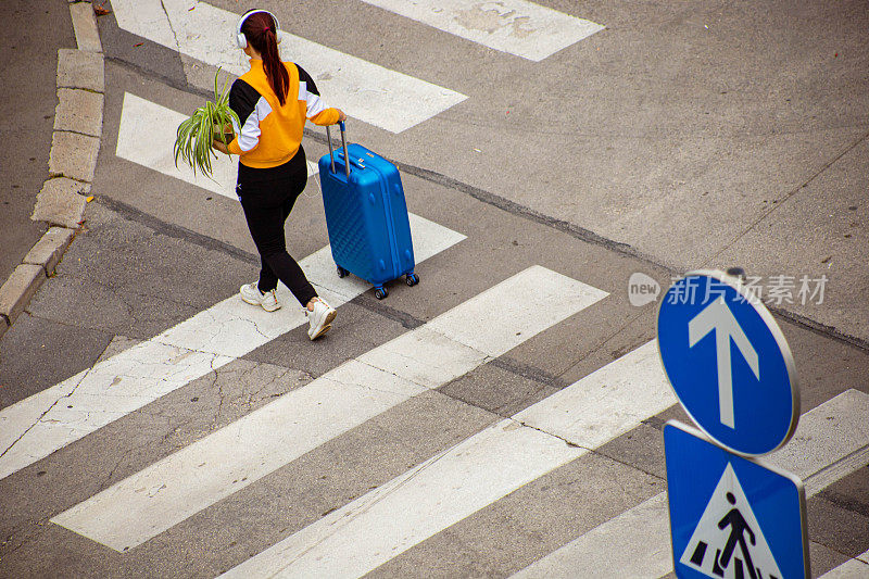 女独自旅行者，推着行李，拿着植物，听着音乐，过马路