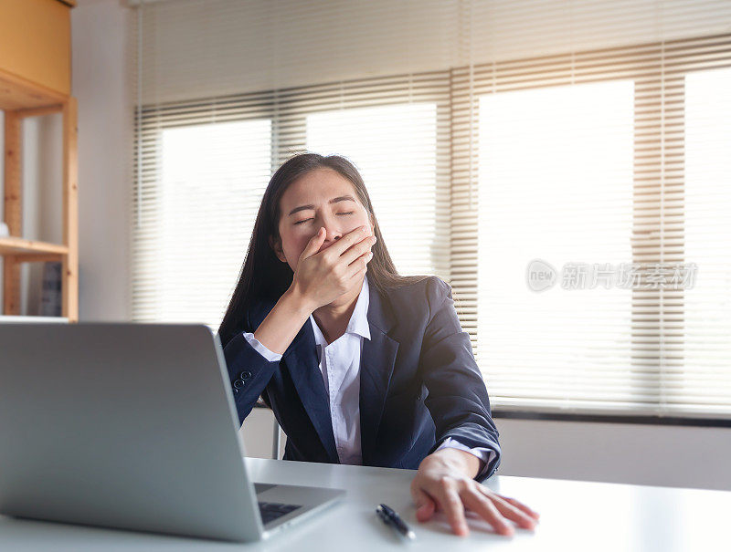 疲倦困倦的商业妇女打哈欠旁边的笔记本电脑，而工作在办公室的桌子，努力工作和过度劳累