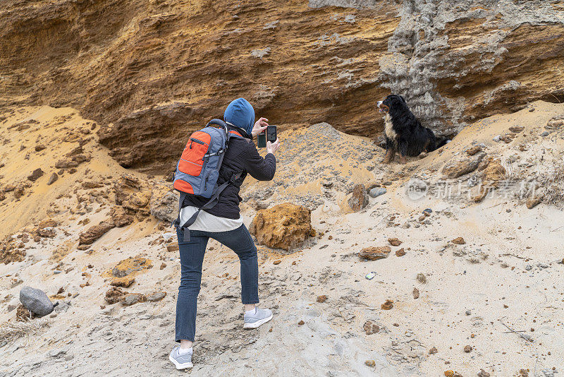 狗主人是一位女士，她戴着蓝色的兜帽，背着背包，正在用智能手机后视镜为她的伯尔尼山狗在沙滩上拍照。