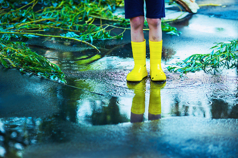夏雨。水坑在沥青。穿着黄色雨靴的孩子。孩子走过水坑。寒冷的秋天的雨。潮湿的天气。水飞溅。倒影在水中