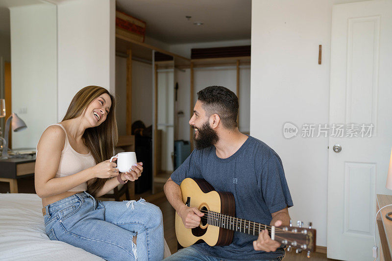 幸福的年轻的多民族夫妇喜欢唱歌和吉他，而坐在卧室在早上。