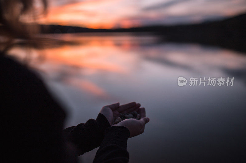 日落时分，一位面无人色的女子站在湖边，手里拿着鹅卵石