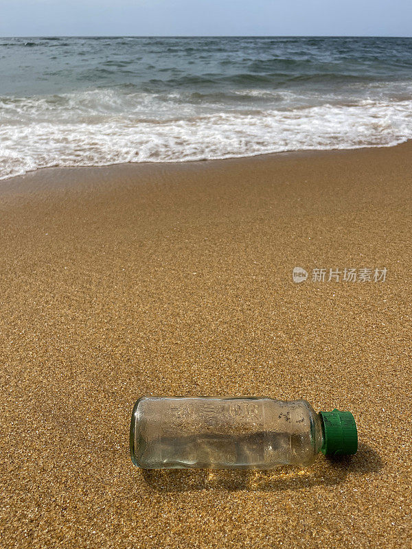 图片:空的，透明的塑料饮料瓶，绿色盖子被冲到退潮水边缘的沙滩上，破碎的海浪冲刷着海岸上的海洋垃圾和污染，乱扔的沙子，肮脏的海滩，关注前景，复制空间