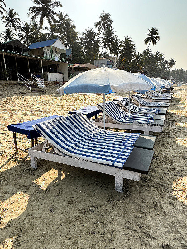 印度果阿，帕洛莱姆海滩，沙滩度假的特写照片，沙滩上有一排排木制的、蓝色软垫的太阳躺床，人们在沙滩上晒日光浴，遮阳伞上有蓝色和白色条纹的毛巾，轮船上有封闭的彩色遮阳伞