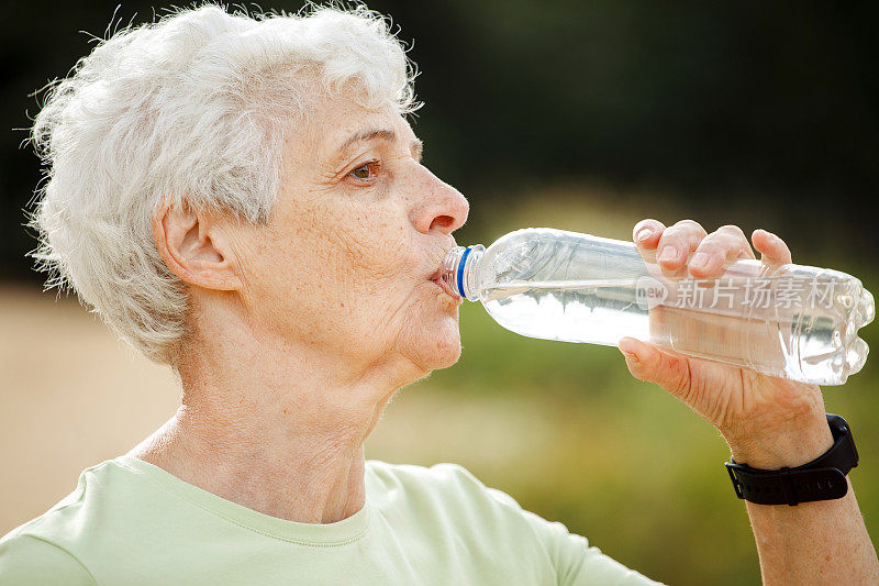 一位灰白短发的老妇人在运动后喝水，在公园里拍照