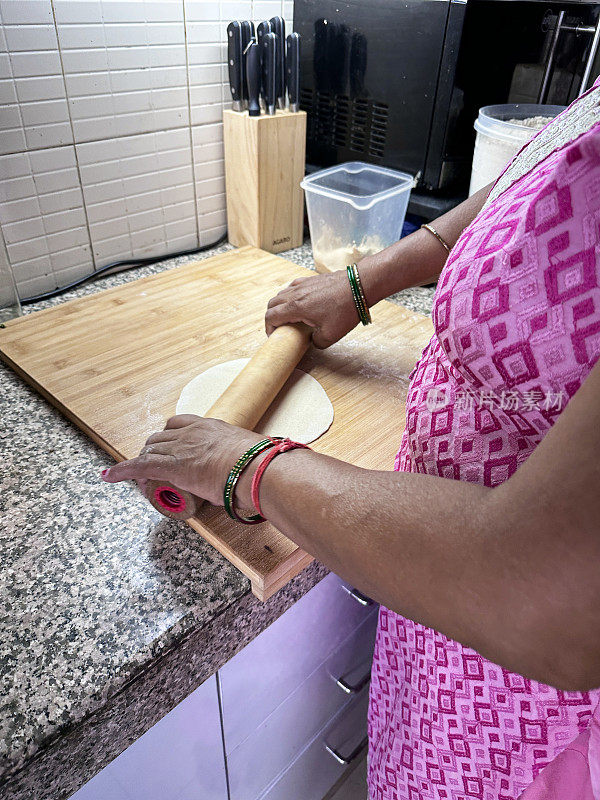 一个面目全非的印度妇女用擀面杖在木制砧板上准备烤面包，厨房柜台的刀架和刀具，全麦atta面粉薄饼面团卷成圆盘，焦点在前景