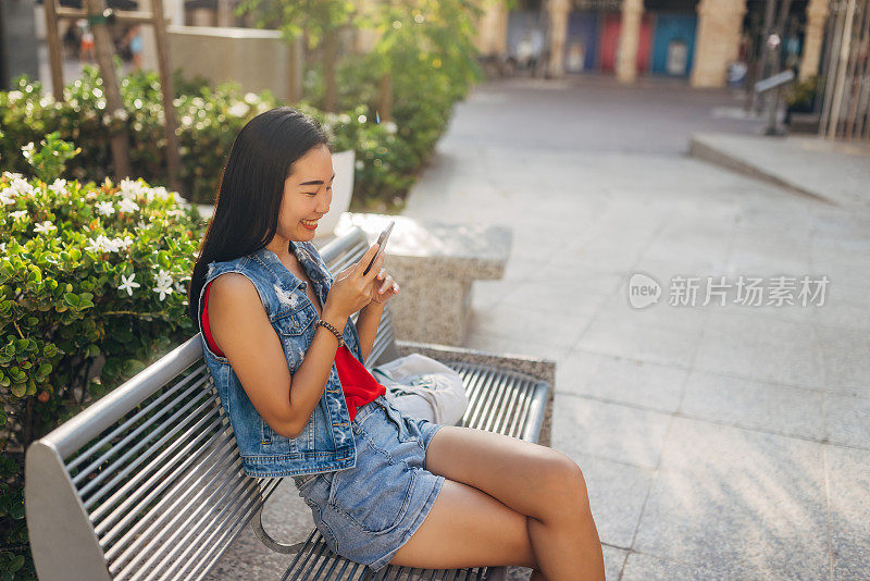 一位无忧无虑的台湾女子坐在公园的长椅上，用智能手机给她的朋友发语音信息