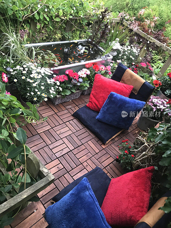 夏天的花园树屋阳台上有红色和蓝色的靠垫，锌池与太阳能喷泉泵，金鱼鱼，边缘植物，红色微型玫瑰，粉色非洲菊花，柚木装饰瓷砖，太阳能灯，照明