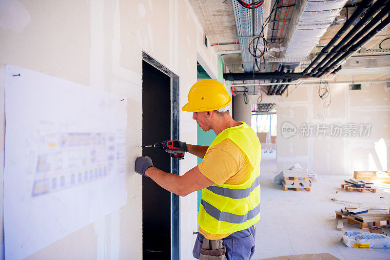 年轻男性建筑工人戴着黄色安全帽，用电钻工具在墙上钻孔。特写前人像与复制空间