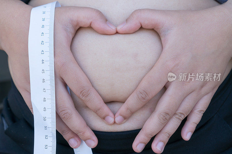 胖女人把心型手放在肚子上进行身体护理，孕期饮食。超重的亚洲年轻女性在过度饮食问题肥胖用卷尺测量，保健美容身体捏她的胖肚子