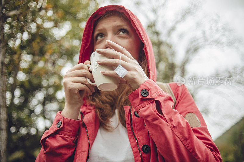 一个年轻的橘黄色头发的女人，穿着红色的雨衣，在雨中走过树林，拿着一杯热茶-放松在一个雨天的自然