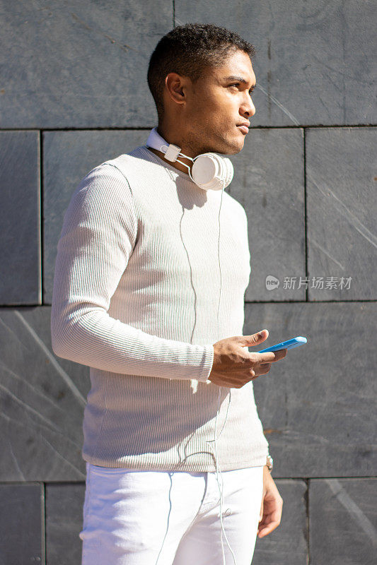 竖直的肖像，严肃的拉丁黑人男子穿着白色连衣裙，看着一边戴着耳机和手机，垂直的墙壁背景