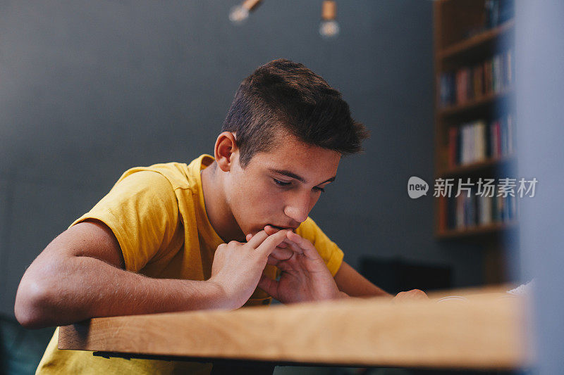 一个小男孩坐在桌旁，手指缠在一起。他靠在他的手，他读他面前的一本书。