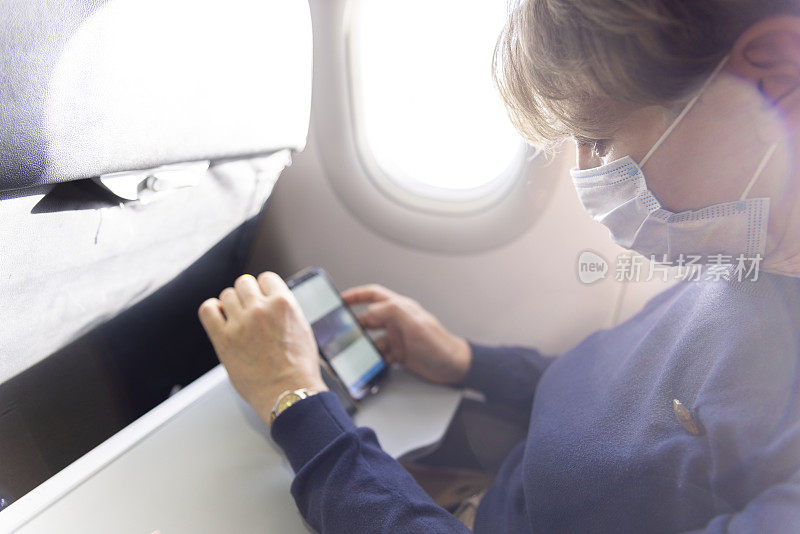 在飞机上看照片。戴着口罩的成熟女性在飞机上用智能手机看照片。