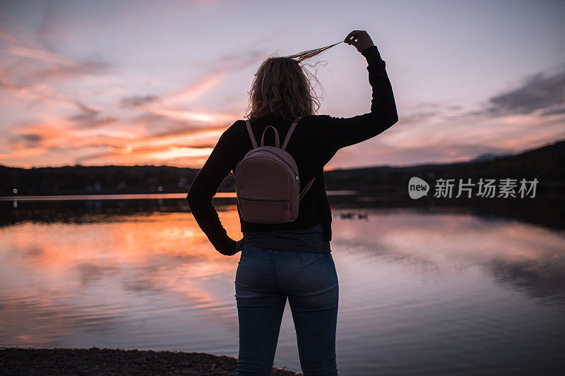 背着双肩包的女人在湖岸欣赏日落，玩她的头发