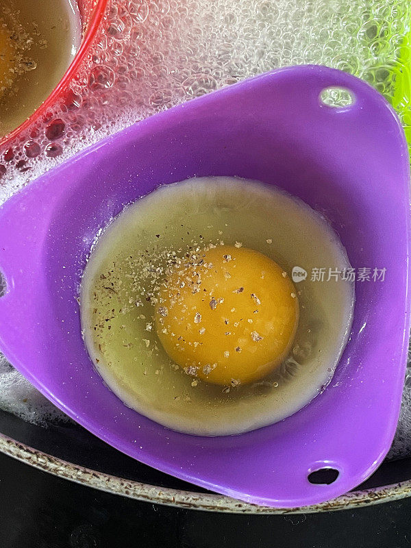 彩色硅胶煮蛋器的全帧图像，在煎锅里煮鸡蛋，紫色煮蛋器在沸水里，鸡蛋用胡椒调味，抬高视图