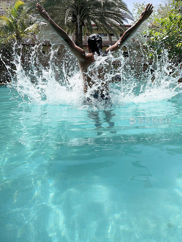 印度男子站在晶莹剔透的水中，扬起溅水的手臂，游泳池，绿松石蓝色马赛克瓷砖，波涛汹涌的水面反射，后视图，重点在前景