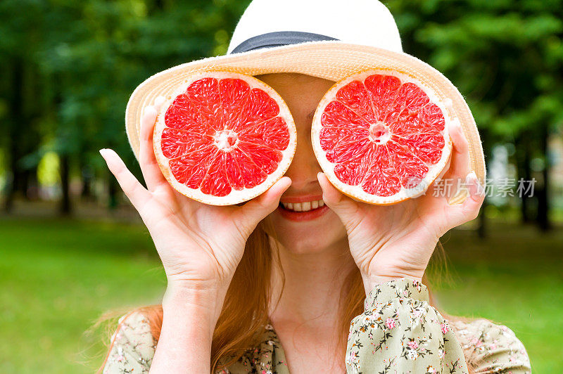 一个年轻美丽的红头发女孩的肖像，拿着一个多汁的葡萄柚靠近她的脸，在一个夏日的公园里享受和微笑。