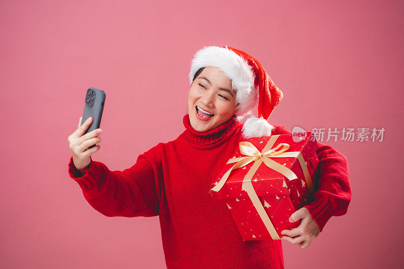 亚洲女人戴着圣诞老人的帽子，拿着礼盒，开心地微笑着，用智能手机在视频会议上说话，孤立在粉红色的背景中。