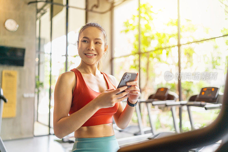 在室内健身俱乐部，一名亚洲女子在阳光下使用智能手机，结束了健身自行车训练