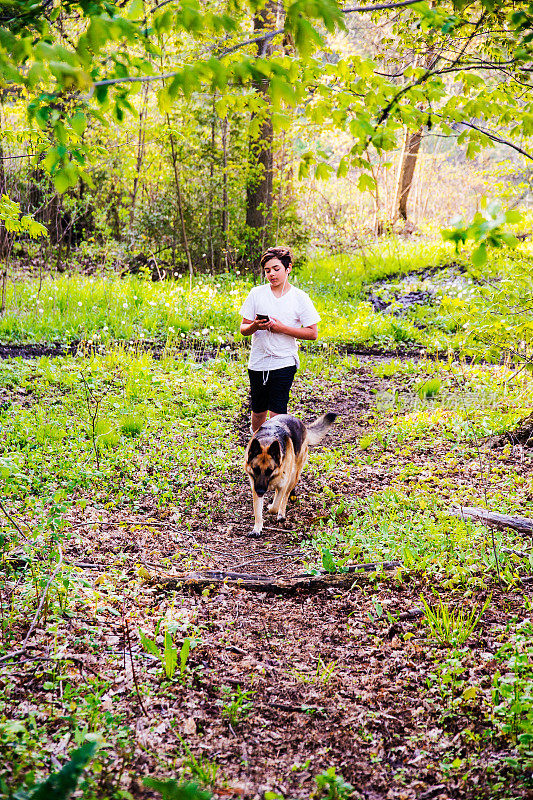 一个十几岁的小男孩带着他的狗在森林里散步，用的是带耳塞的电话。