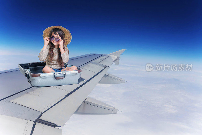 带着草帽和眼镜微笑的小女孩坐在飞机机翼上的行李箱里