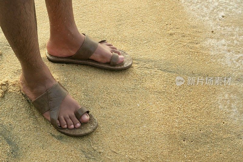 这张照片拍摄于印度南部喀拉拉邦的Kollam，照片中，印度男子穿着棕色皮凉鞋在金色的沙滩上行走，男子湿漉漉的毛腿和棕色人字拖凉鞋在海边被海浪拍打，赤脚走在海滩上