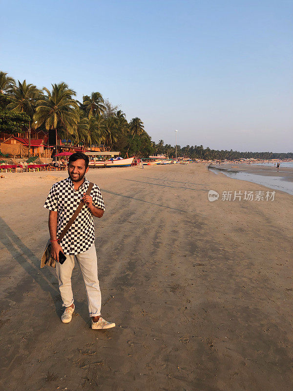 这是一个年轻的印度男子在海滩度假时，穿着黑白格子衬衫和蓝色牛仔裤子，走在海边的照片。一个英俊的印度男子穿着鞋子在喀拉拉邦果阿海边，微笑着走在海滩上