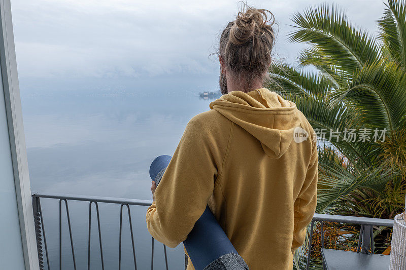 一个年轻人拿着瑜伽垫在她的阳台上沉思着湖和山的景色