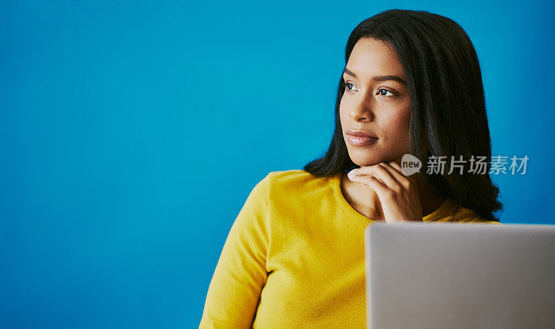 剪裁照片的一个有吸引力的年轻女子看起来深思熟虑，而坐在她的办公桌在工作室的蓝色背景