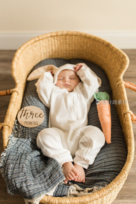一个16周大的兔宝宝穿着白色的兔宝宝服装，躺在一个舒适的海草摩西篮子里和他的胡萝卜睡觉