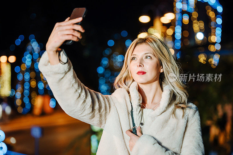 快乐的年轻女子在夜晚的城市户外用智能手机自拍