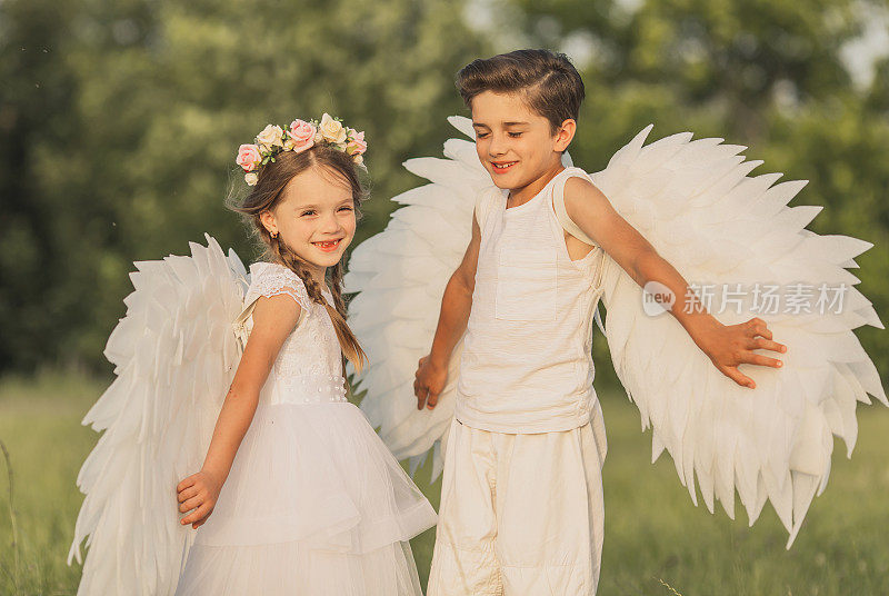 两个白翅膀的天使在绿色的草地上。金发女孩在裙子和深色头发的男孩在夏天的日落背景
