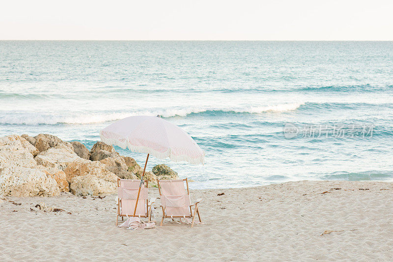 波西米亚条纹粉白色沙滩伞和折叠沙滩椅在棕榈海滩，佛罗里达州日落时分
