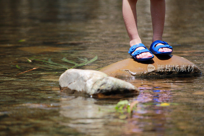 亚洲男婴穿着拖鞋站在小溪里的一块石头上