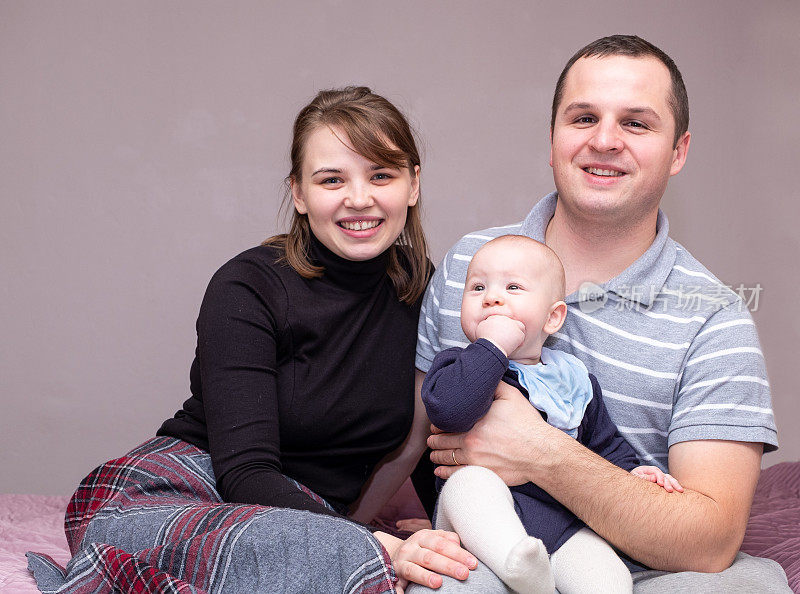 这是一个年轻的白人家庭，一对年轻的男人和女人抱着一个婴儿，他们看着镜头，微笑着。