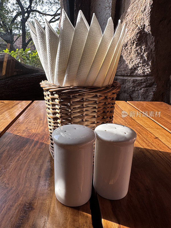 白色的盐和胡椒罐的特写图像，柳条篮子餐巾架，在木制餐厅的桌子上，露天用餐区，重点在前景