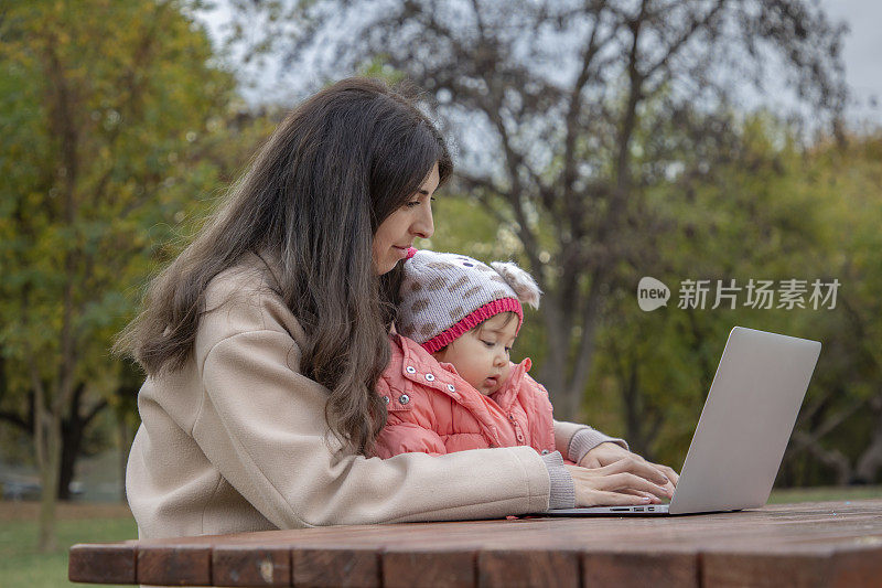 一个女人在大自然中摆弄着她的电脑，她的孩子坐在她的腿上。