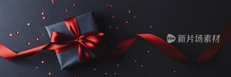 礼品盒与红色蝴蝶结黑色背景与红色五彩纸屑。圣诞节、生日、周年纪念的概念。俯视图，平铺