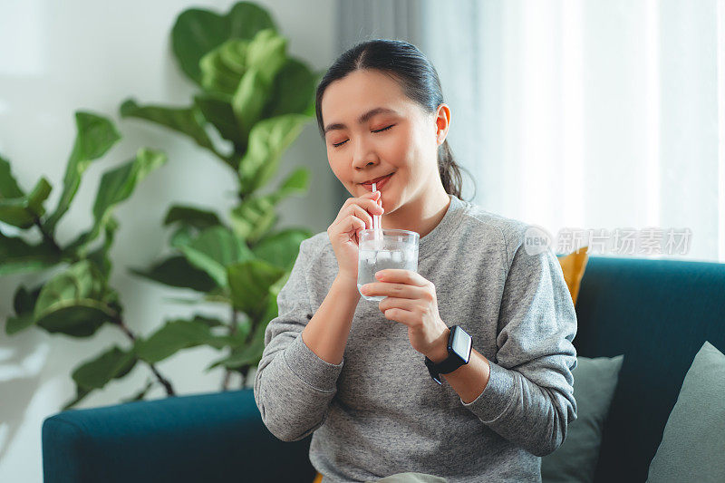 亚洲妇女坐在客厅的沙发上用吸管喝凉水休息。