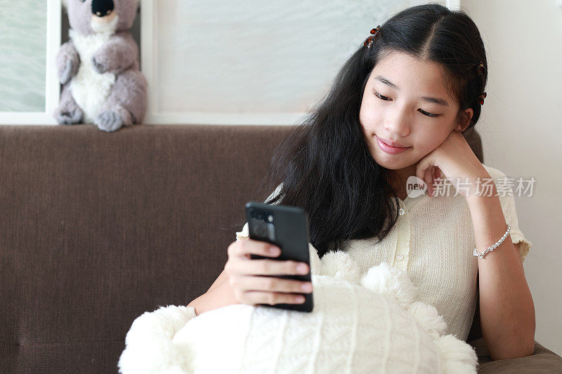 一个无忧无虑、放松的年轻亚洲女孩坐在沙发上玩她的智能手机。她正在浏览社交媒体、浏览互联网、网上购物或与朋友聊天。