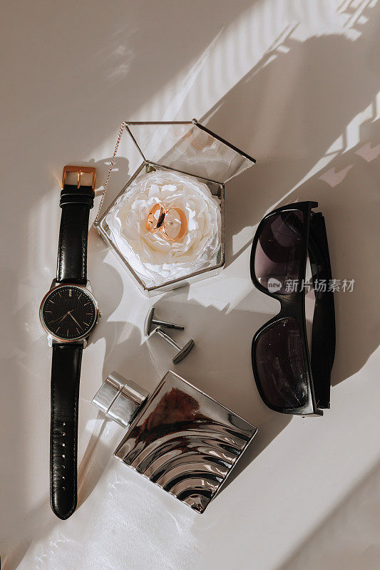 新郎的细节放在一张白色的桌子上。黑眼镜，手表，金婚戒，男士香水，袖扣