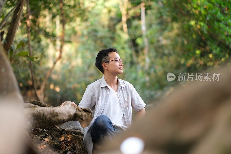 年轻的亚洲人坐在岩石，溪流和森林中的岩石上，仰望大自然，周围有树木和灯光照耀的背景，带着淡淡的微笑和清新的感觉，人们对自然负责的观念。