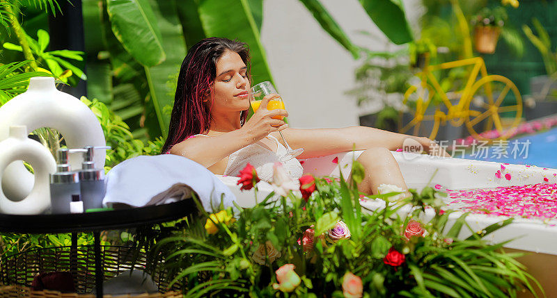 印度微笑的z世代女性坐在浴缸里放松，喝着健康的新鲜果汁。美丽的年轻成年女孩早上沐浴玫瑰花瓣水享受暑假在酒店度假别墅之旅