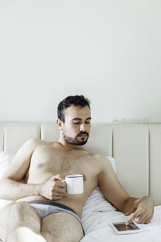 年轻人躺在床上，一边喝着咖啡一边用着平板电脑。