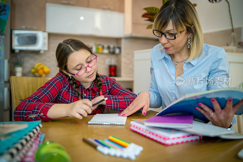 迷人的年轻女子和她可爱的小女儿正坐在桌旁一起做作业。母亲帮助女儿学习学校课程。