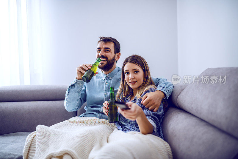 一对漂亮的白人夫妇盖着毯子坐在客厅里看电视喝啤酒。拿着遥控器换频道的女人。