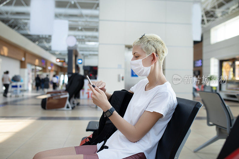 年轻的女旅行者戴着面具用手机和坐在机场的距离